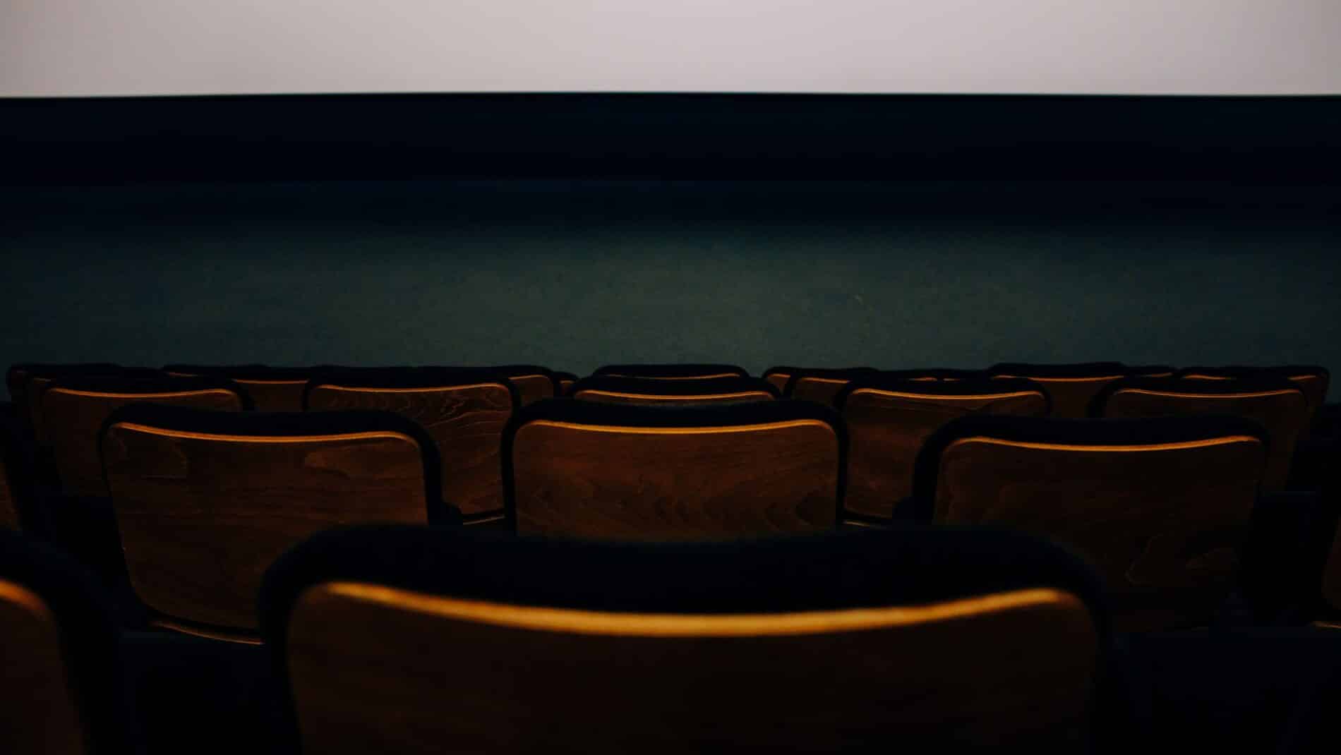 An empty auditorium with wooden seats facing a blank theater screen.