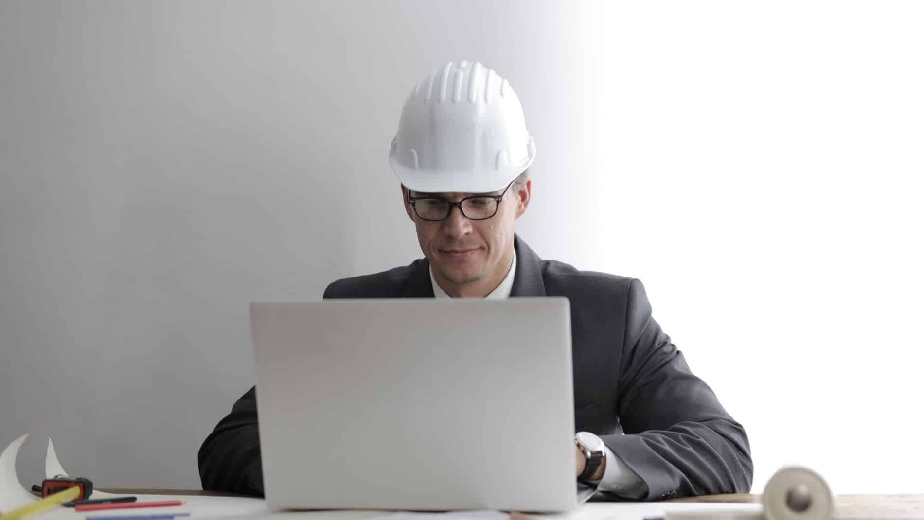 Serious concentrated engineer in hardhat and formally suit sitting at table with blueprints while working on laptop against gray background