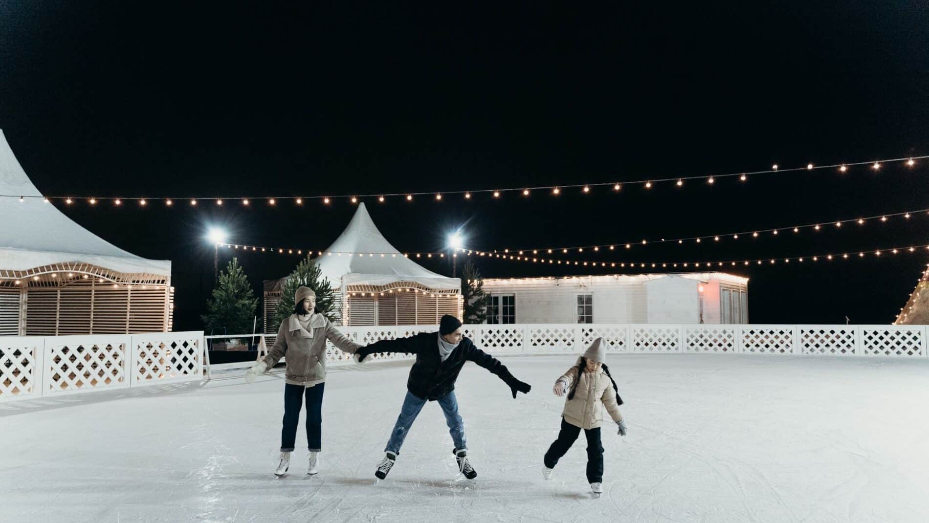 A family enjoying ice skating together on an outdoor rink at night under festive string lights.
